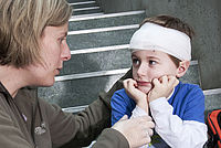 Das Bild zeigt einen kleinen Jungen, der auf einer Treppe sitzt. Sein Kopf ist mit einem Verband verbunden. Neben ihm sitzt eine Frau, die ihn tröstet. Sie legt eine Hand auf seine Schulter und schaut ihn liebevoll an.