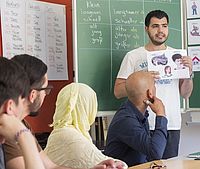 Auf dem Bild ist eine Person zu sehen, die vor der Klasse steht und den interessierten Flüchtlingen die deutsche Sprache beibringt. Hinter ihm ist eine beschriebene Schultafel zu sehen.