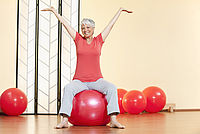 Das Bild zeigt eine Frau in einem Sportstudio, die auf einem großen, runden Pezziball sitzt. Sie hat beide Hände über den Kopf gehoben und lächelt freundlich in die Kamera. Im Hintergrund sind weitere Pezzibälle sichtbar, die im Raum verteilt sind.