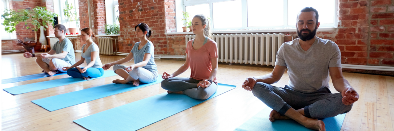 Mehrere Personen sitzen auf Yogamatten in einem hellen Raum mit Backsteinwänden und großen Fenstern und meditieren mit geschlossenen Augen und gekreuzten Beinen.