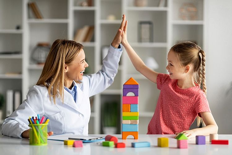 Auf dem Bild sind eine Frau und ein Mädchen zu sehen, di sich in einem Schulraum befinden. Die beiden haben gerade eine Würfelaufgabe gelöst. Die Frau gibt dem Mädchen fünf, es ist eine Geste des Erfolgs. Beide lächeln sich freundlich an und scheinen sich über ihre gemeinsame Leistung zu freuen.
