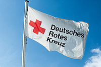Das Bild zeigt eine weise Fahne mit dem Logo des Deutschen Roten Kreuzes (DRK) darauf. Im Hintergrund ist ein blauer Himmel zu sehen.