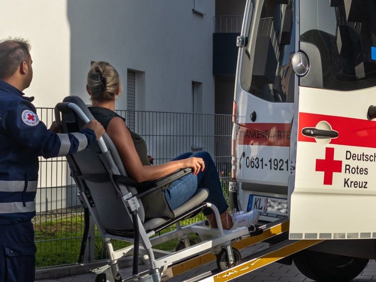 Auf dem Bild sind ein Krankenfahrzeug und ein Mitarbeiter in Dienstkleidung zu sehen. Der Mitarbeiter schiebt eine junge Frau im Rollstuhl die Rampe des Fahrzeugs hinauf, um sie in das Fahrzeug zu bringen.