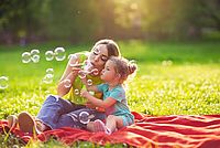Auf dem Bild sieht man eine junge Mutter, die mit ihrem kleinen Mädchen auf einer grünen Wiese sitzt. Beide genießen gemeinsam die schöne Natur und machen Luftblasen.