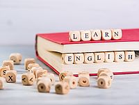 Ein aufgeschlagenes Buch mit rotem Einband liegt auf einem Tisch. Darauf und davor befinden sich kleine Holzwürfel mit Buchstaben. Aus diesen Würfeln sind die Wörter „LEARN“ (oben auf dem Buch) und „ENGLISH“ (vor dem Buch) gelegt. Im Vordergrund liegen weitere Buchstabenwürfel verstreut.