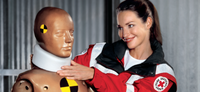 Auf dem Bild sind zwei Personen zu sehen. Eine ist eine Rettungssanitäterin, die in Berufskleidung gekleidet ist. Sie schaut freundlich zur Dummy-Puppe hin, die neben ihr steht. Die Dummy-Puppe wird für Erste-Hilfe-Übungen verwendet. Das Foto ist ein Portrait, das die Sanitäterin und die Dummy-Puppe zeigt.