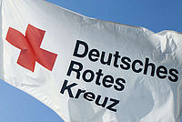 Das Bild zeigt eine weise Fahne mit dem Logo des Deutschen Roten Kreuzes (DRK) darauf. Im Hintergrund ist ein blauer Himmel zu sehen.