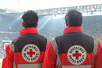 Auf dem Bild sind zwei Sanitäter in Berufskleidung zu sehen, die mit dem Rücken zur Kamera stehen. Sie befinden sich auf einem Fußballstadion während eines Spiels. Die Sanitäter sind in der Nähe des Spielfelds und scheinen das Spiel zu beobachten.