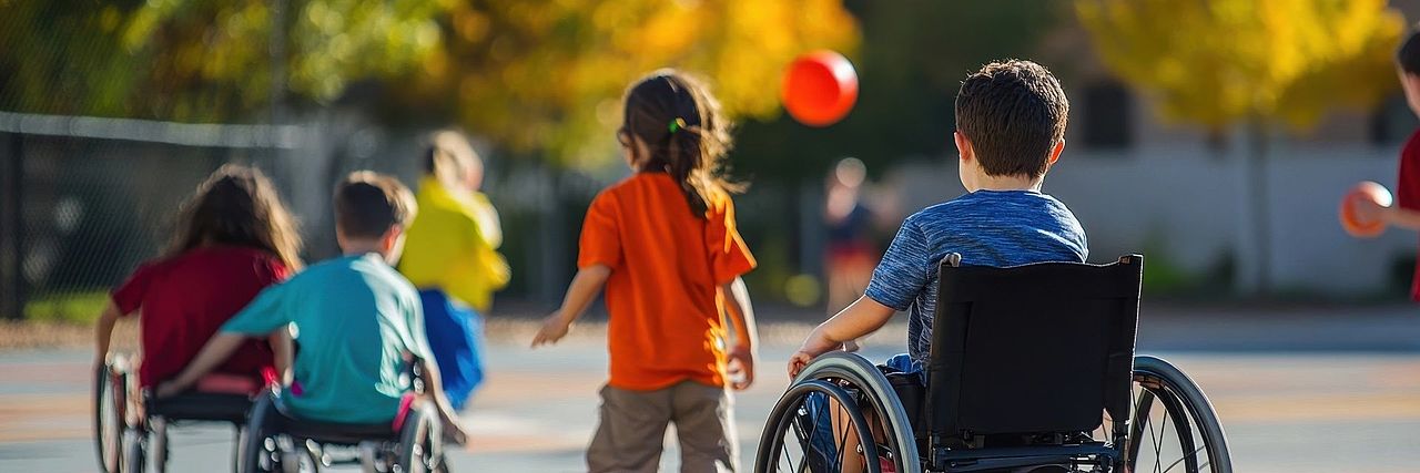 Auf dem Schulhof spielen fünf Kinder mit Bällen. Drei der Kinder sitzen im Rollstuhl, alle sind mit dem Rücken zur Kamera. Sie haben Spaß beim gemeinsamen Spiel und zeigen, dass Freude und Bewegung für alle möglich sind.