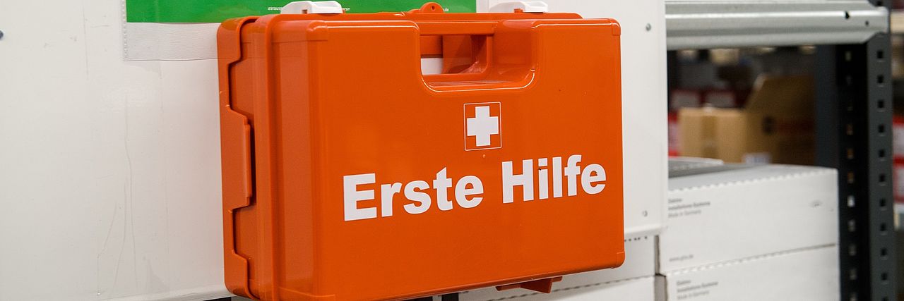 Auf dem Bild ist ein Verbandkasten in Farbe orange und weisem Text "Erste Hilfe" zu sehen.hen. Ein Wort Erste Hilfe