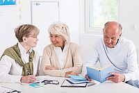 Auf dem Bild sind zwei Frauen mittleren Alters und ein älterer Mann zu sehen. Sie sitzen gemeinsam an einem Tisch in einem Schulungsraum. Vor ihnen liegen Übungshilfen und Bücher. Die drei Personen scheinen sich freundlich zu unterhalten.