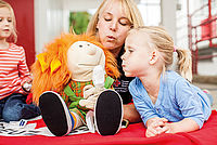 Auf dem Bild sind eine Kindergärtnerin und zwei Vorschulkinder zu sehen. Alle drei sitzen zusammen und spielen mit  Stoffpuppe Paula..