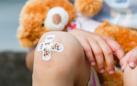 Ein Kind sitzt mit einem kleinen Stoffbär auf dem Schoss. Auf dem Knie des Kindes klebt ein buntes Kinderpflaster.