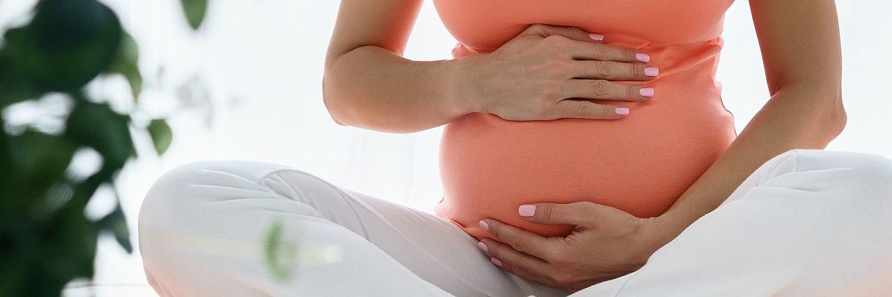 Schwangere Frau sitzt im Schneidersitz auf dem Boden und hält mit beiden Händen ihren Babybauch.