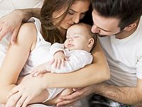 Auf dem Bild sind eine junge Frau und ein junger Mann zu sehen. Der junge Mann umarmt die junge Frau liebevoll. In der Mitte zwischen ihnen liegt ein Neugeborenes, das liebevoll gehalten wird.