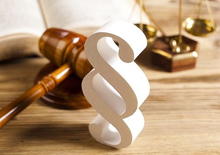 Das Bild zeigt drei symbolträchtige Objekte aus dem juristischen Bereich auf einem Holztisch: Im Vordergrund steht ein großes, plastisches Paragraphenzeichen (§) in Weiß. Links dahinter liegt ein hölzerner Richterhammer auf einem runden Holzblock. Im Hintergrund ist eine goldene Justitia-Waage zu erkennen