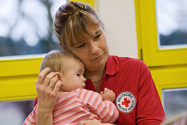 Eine Frau in DRK Kleidung hält liebevoll ein zweijähriges Mädchen im Arm.