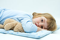 Auf dem Bild liegt ein Kind auf einer Kuscheldecke, das ein Kuscheltier in den Armen hält. Das Kind schaut traurig und müde in die Kamera, weil es krank ist.