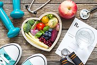 Auf einem Holztisch liegen blaue Hanteln, Sportschuhe, ein Stethoskop, ein roter Apfel, ein Rezeptblock sowie ein Handtrainer. Im Zentrum steht eine herzförmige Schale, gefüllt mit buntem Obst und Gemüse wie Brokkoli, Tomate, Banane, Heidelbeeren, Granatapfel und mehr – Symbol für gesunde Ernährung.