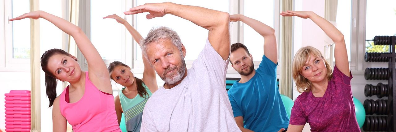 Zwei Männer und drei Frauen über 50 sitzen im Schneidersitz auf Yogamatten in einem Übungsraum und machen gemeinsam eine Yoga-Übung.