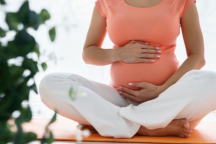 Schwangere Frau sitzt im Schneidersitz auf dem Boden und hält mit beiden Händen ihren Babybauch