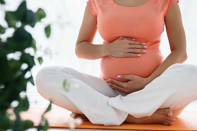Schwangere Frau sitzt im Schneidersitz auf dem Boden und hält mit beiden Händen ihren Babybauch